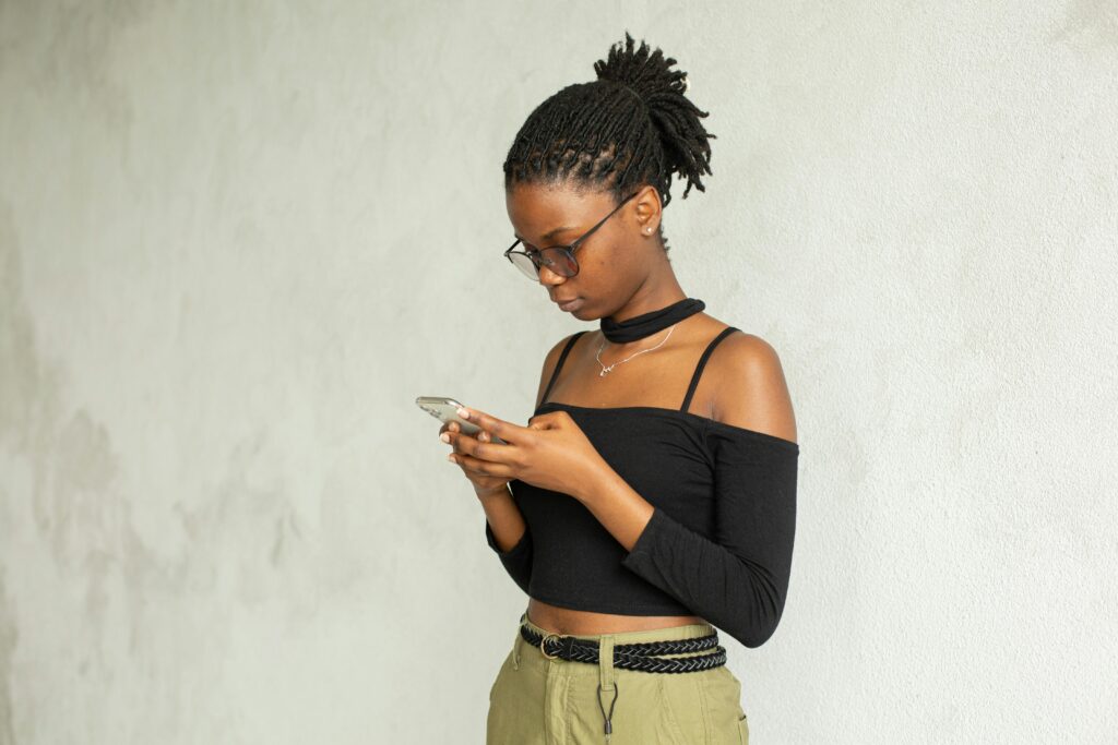 Young African American female in casual clothes and eyeglasses standing against white wall and browsing mobile phone